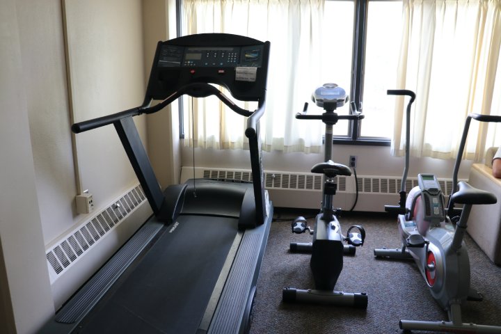 The image shows a small exercise room with a treadmill and two stationary bikes positioned near a window. The equipment is arranged closely, with light streaming through partially drawn curtains, creating a cozy and compact space for indoor exercise.